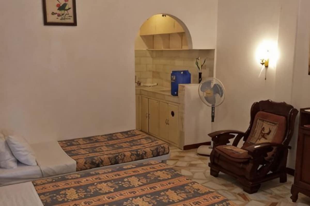 A room with two beds, a wooden chair, a fan, and a kitchen area with an archway.