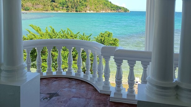Un balcon abrité, doté de balustrades blanches, qui offre une vue sur une mer turquoise et une colline verdoyante.