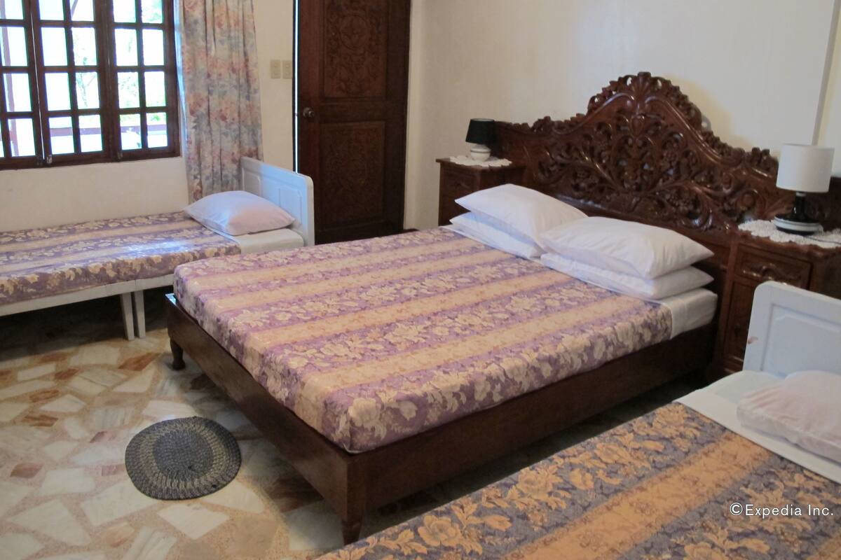 Une chambre avec deux lits simples, une tête de lit en bois, une table de chevet avec une lampe et une grande fenêtre avec des rideaux.