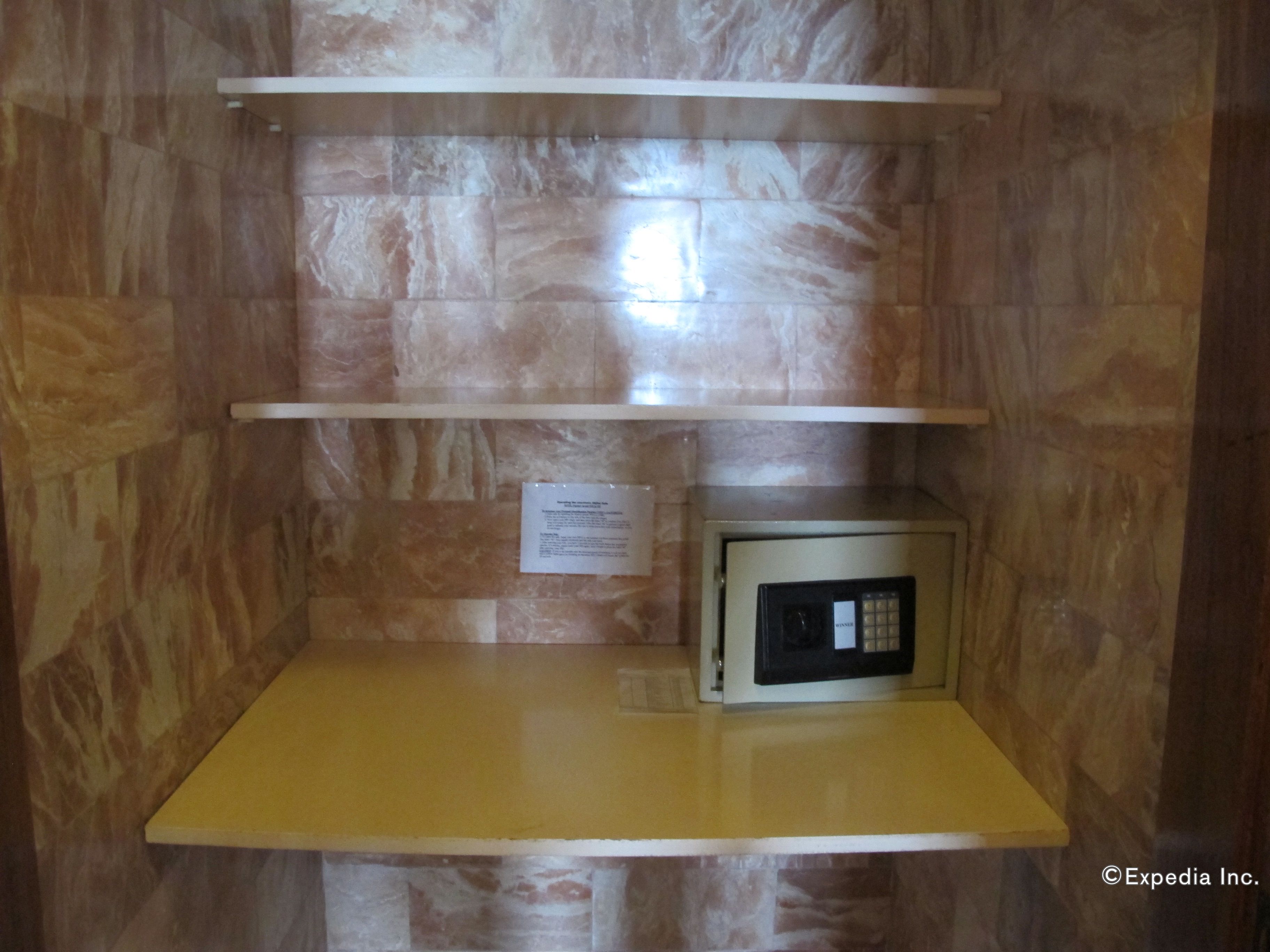 A small kitchenette with a microwave on a yellow countertop, marble walls, and a white shelf above.