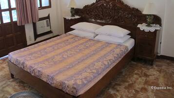 A bedroom with a large bed, two bedside tables with lamps, a wooden headboard, a window with curtains, and a radiator.