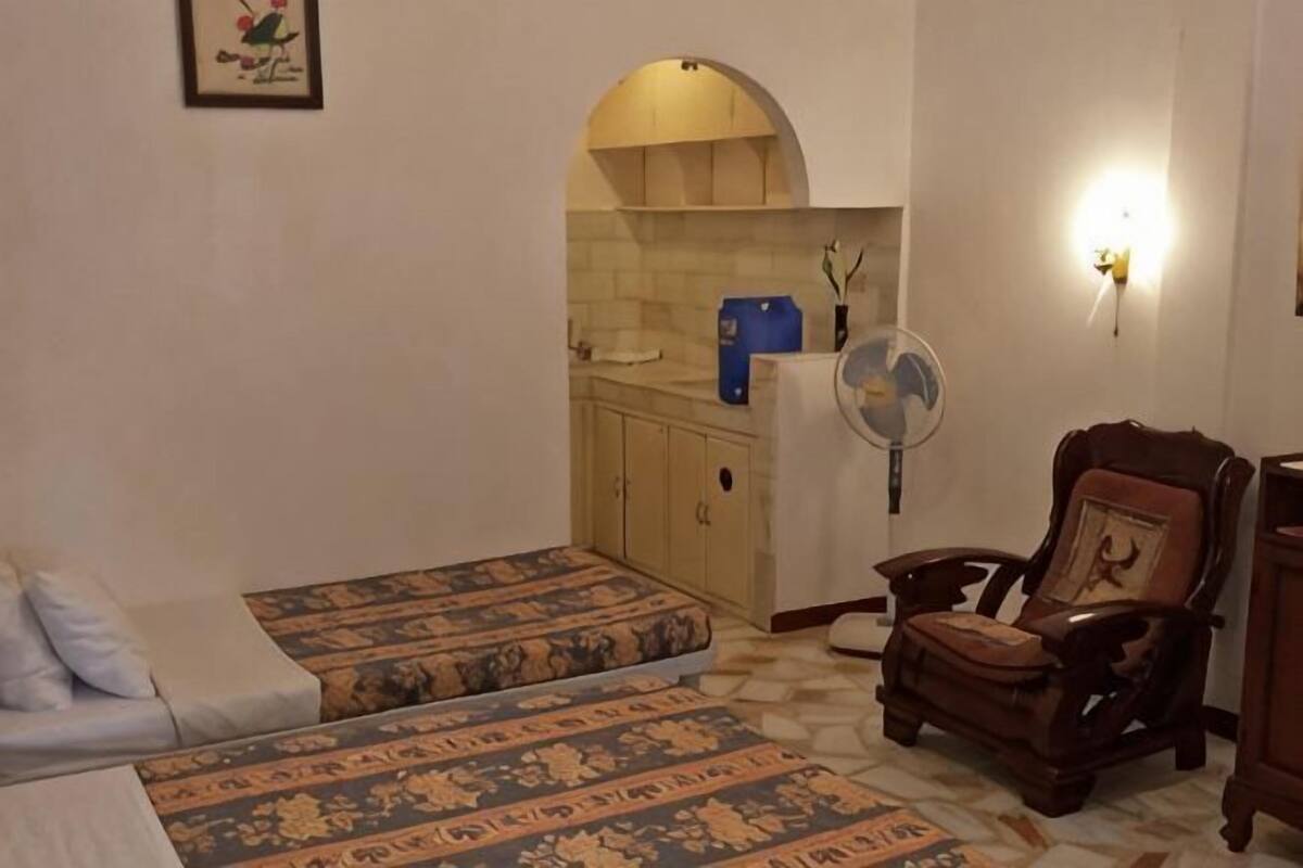 Une chambre avec deux lits, une chaise en bois, un ventilateur et un coin cuisine séparé par une arche.