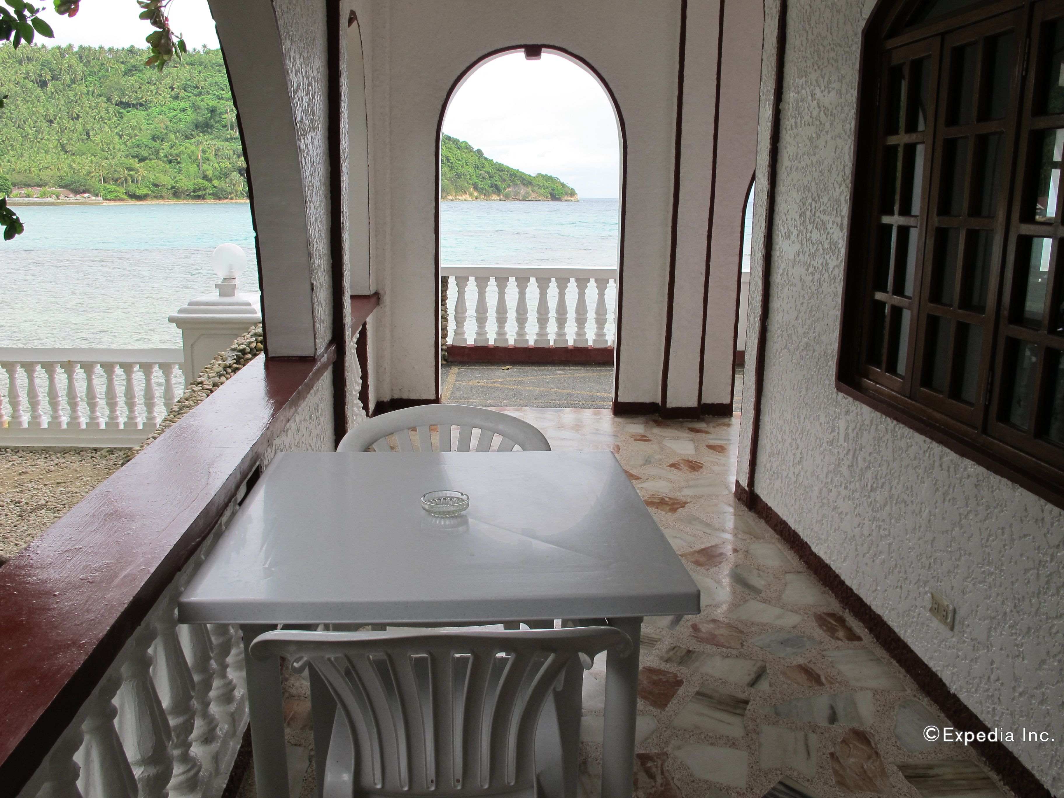 Un balcón con una mesa y sillas blancas, con vistas al agua y una ladera verde.