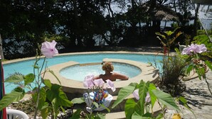 Un bain à remous entouré de verdure et une piscine, avec une chaise et une serviette à proximité.