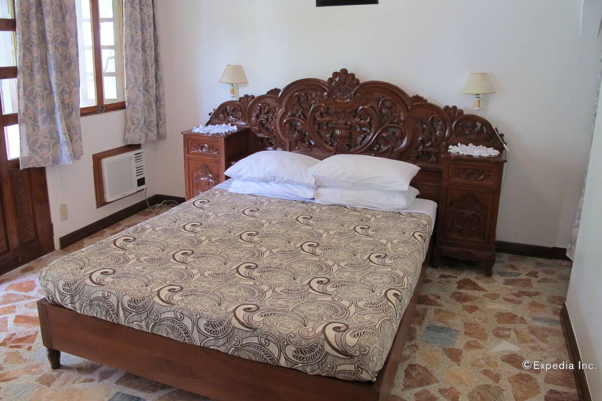 Une chambre à coucher avec un grand lit, une tête de lit en bois, deux tables de chevet, une commode, une fenêtre avec des rideaux et un tableau accroché au mur.