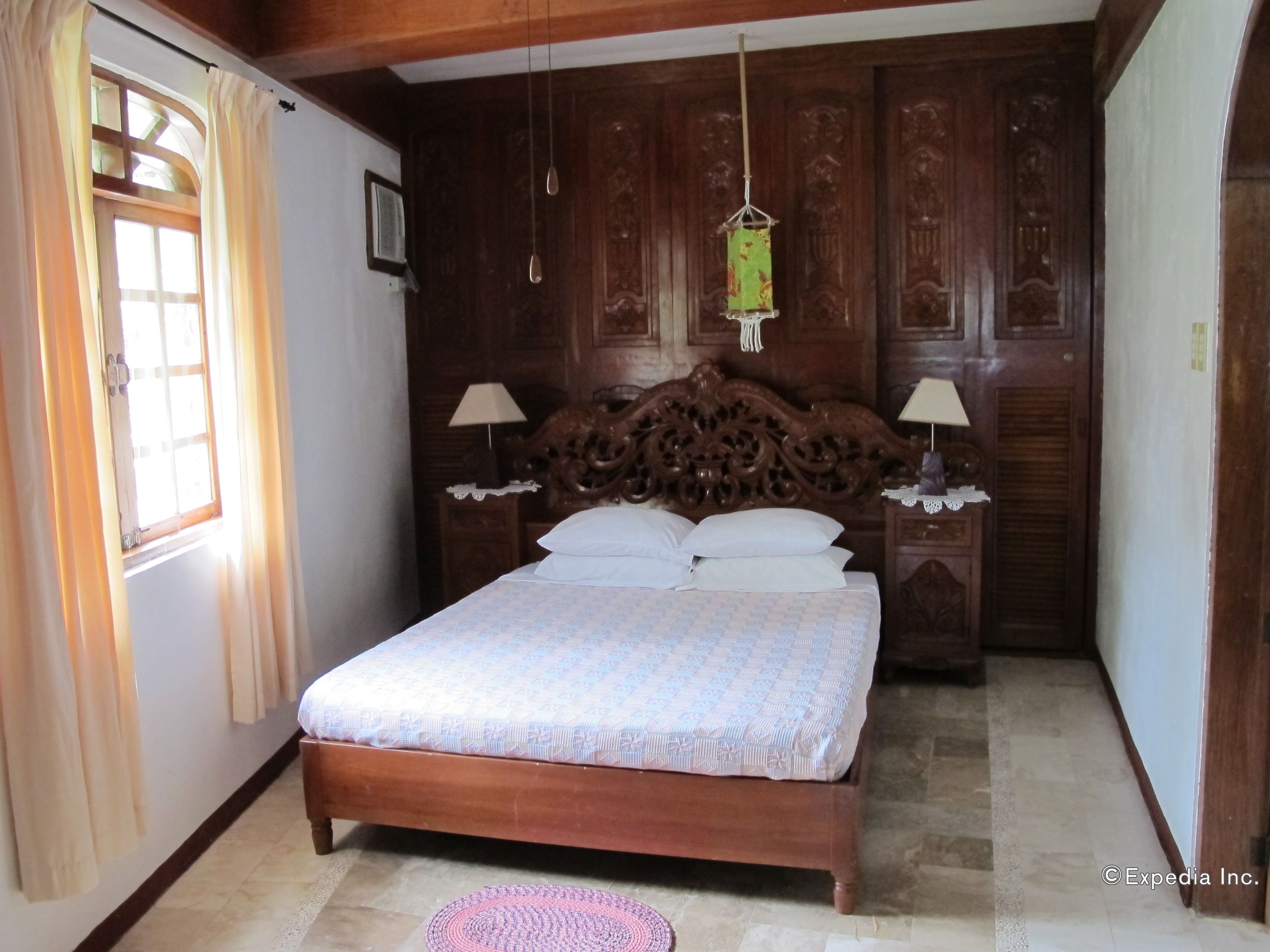 Quarto com cama de madeira, duas mesas de cabeceira com luminárias, uma janela com cortinas e um lustre.