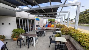 Ein Terrassenbereich im Freien mit Tischen und Stühlen, im Hintergrund ein Spielbereich für Kinder und ein Gebäude mit großen Fenstern.