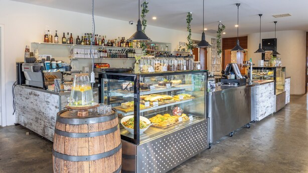 Eine Bar mit einer Theke, Regalen voller Flaschen, einer Kühltheke mit Speisen und hängenden Pflanzen.