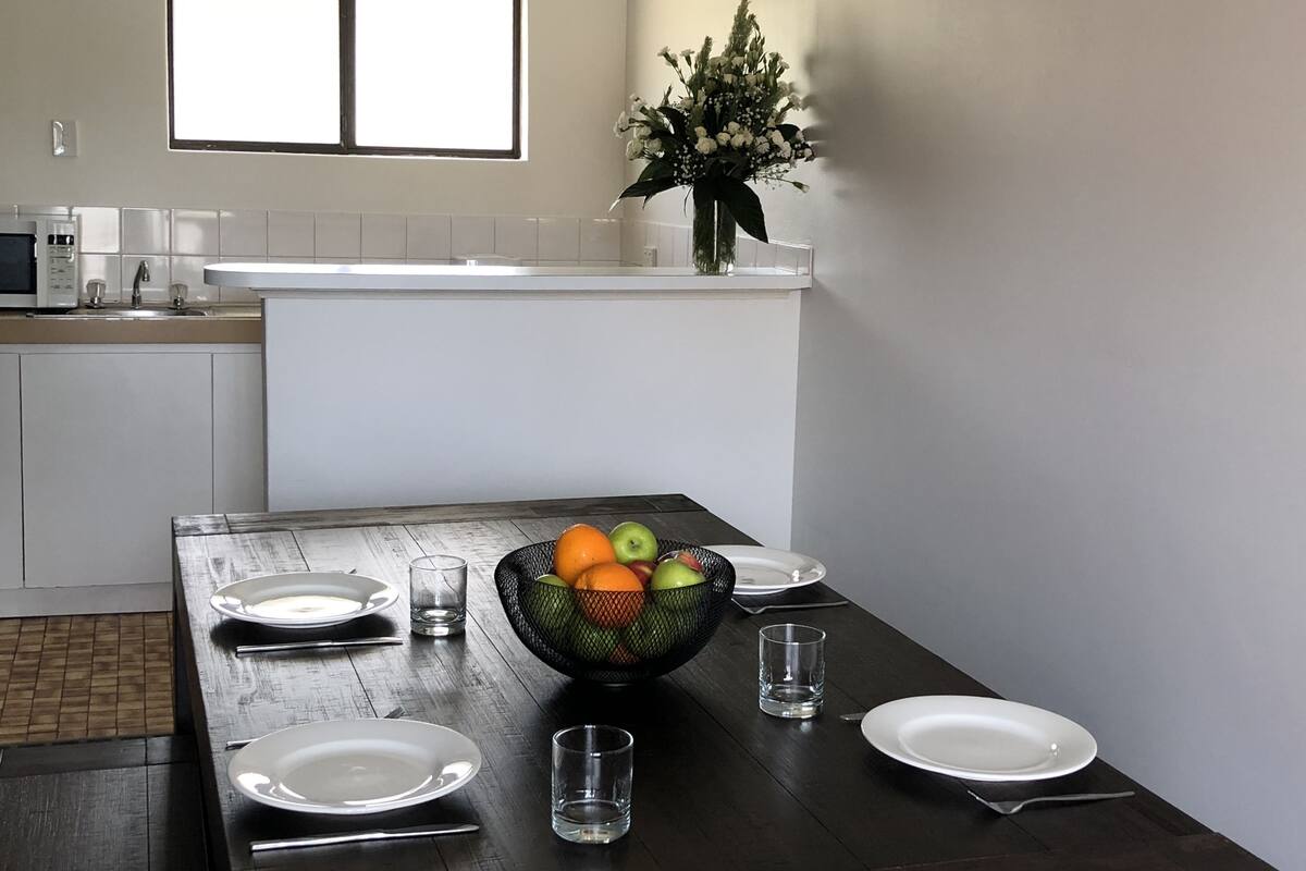 A modern kitchen with white cabinets, a white countertop, and a dining table set with plates, glasses, and a fruit bowl.