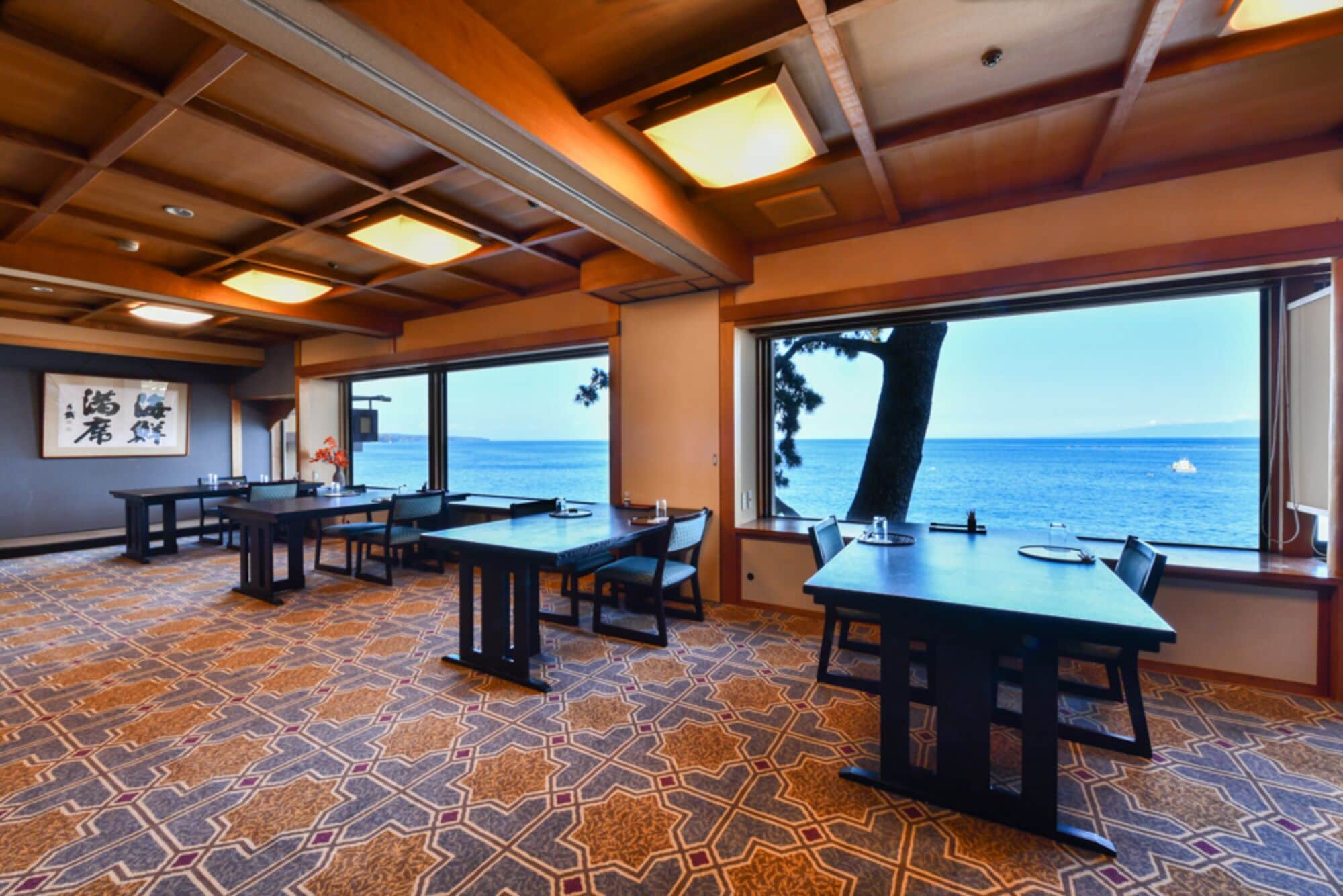 A restaurant with tables and chairs, large windows with a view of the sea, and patterned floor tiles.