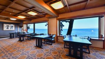 A restaurant with tables and chairs, large windows with a view of the sea, and patterned floor tiles.