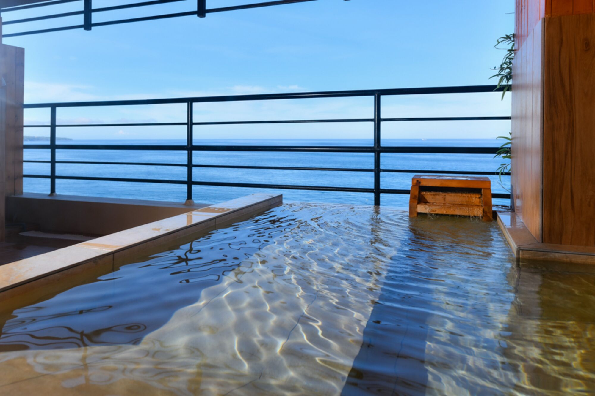 An indoor pool with clear water, a wooden bench, and a view of the sea through a metal railing.