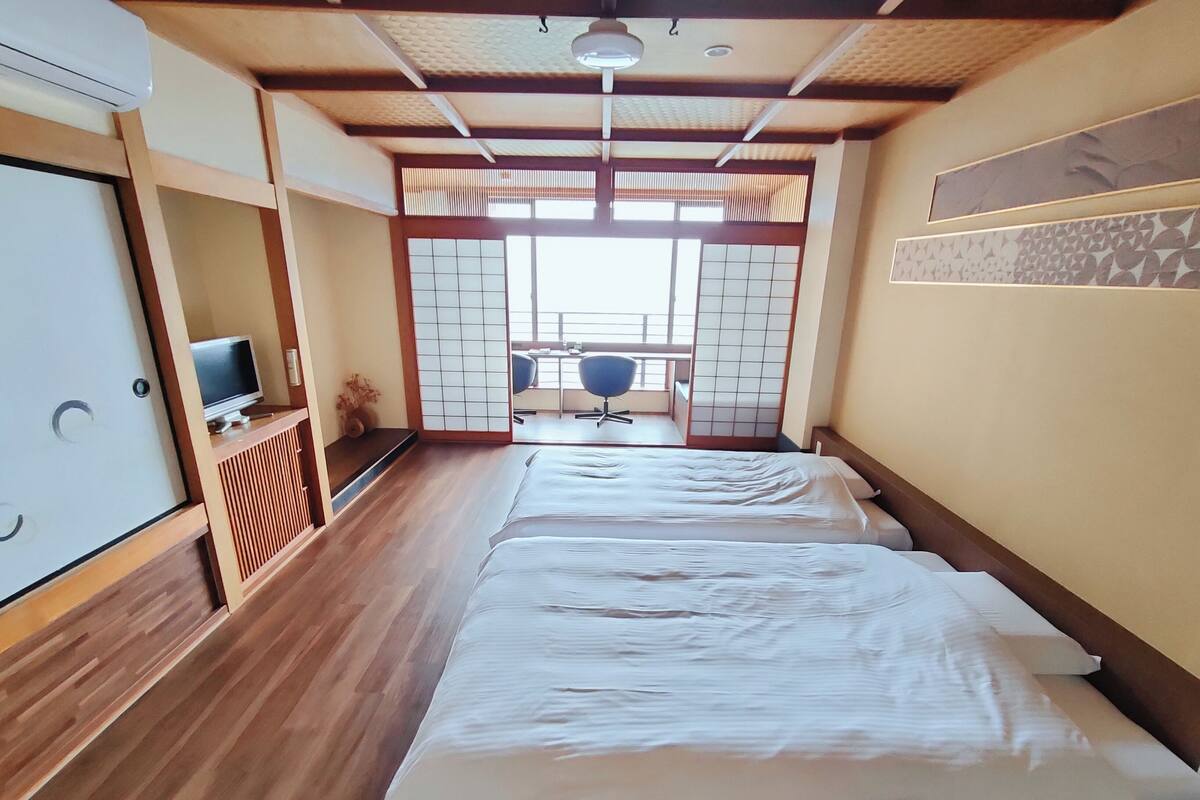 Une chambre d’hôtel avec un plancher en bois, un lit avec une literie blanche, un téléviseur posé sur un meuble et une porte coulissante donnant sur un balcon équipé d’une chaise.