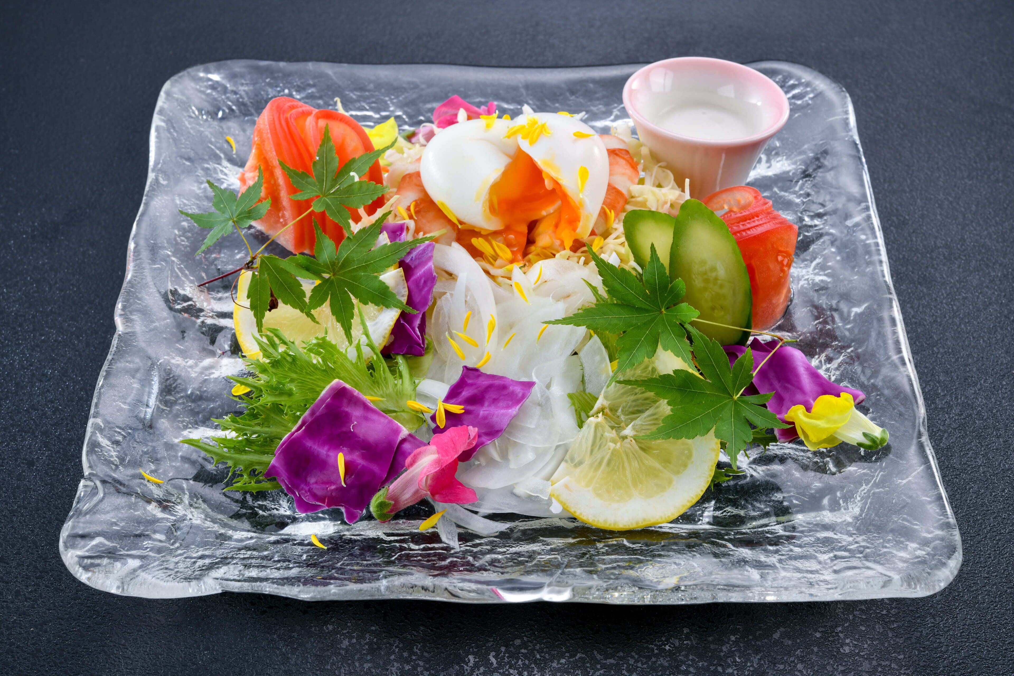 A clear glass dish with a salad, including sliced vegetables and a poached egg, on a dark surface.