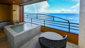 A spa area with a large bathtub, a small table, and a wicker chair, overlooking a body of water with a railing.