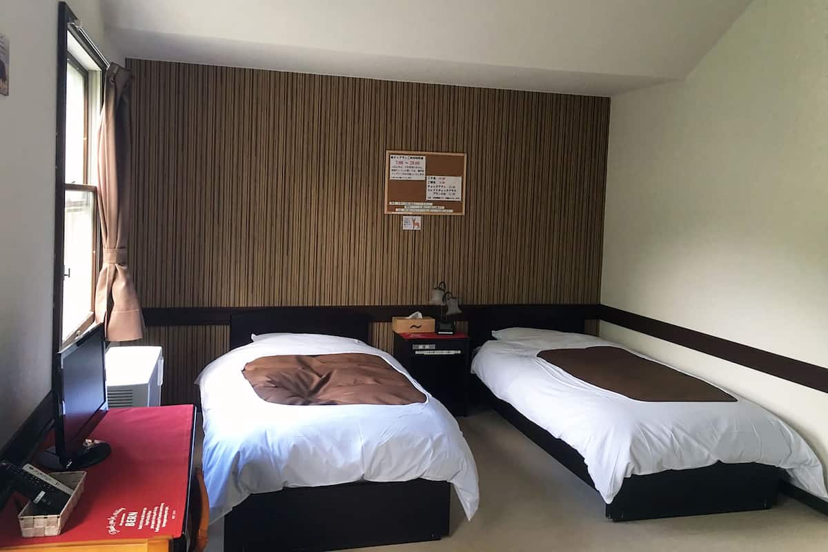 A hotel room with two beds, a desk, a chair, a red table, a bulletin board, and a window with curtains.