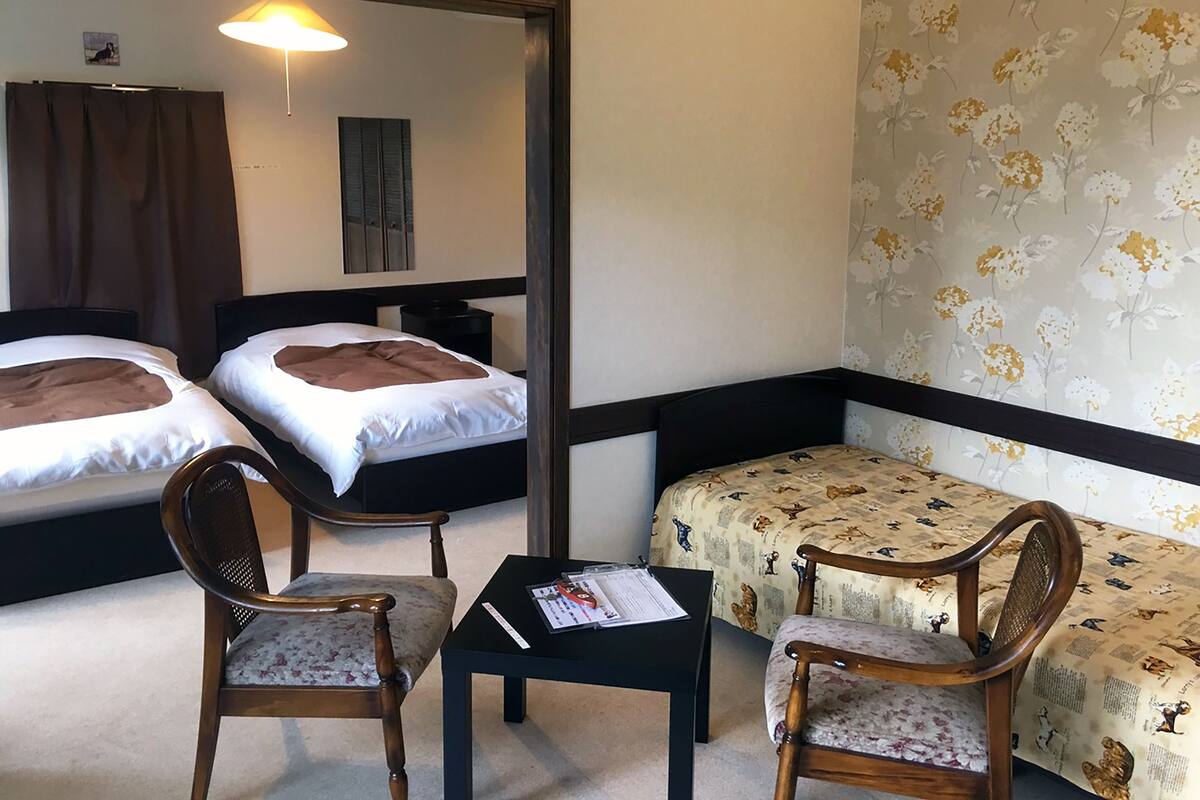 A hotel room with two beds, a small table, two chairs, a mirror, and floral wallpaper.