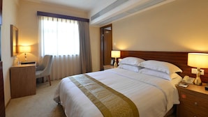 A hotel room with a large bed, a desk, a chair, a desk lamp, a bedside table with a lamp, and a window with curtains.
