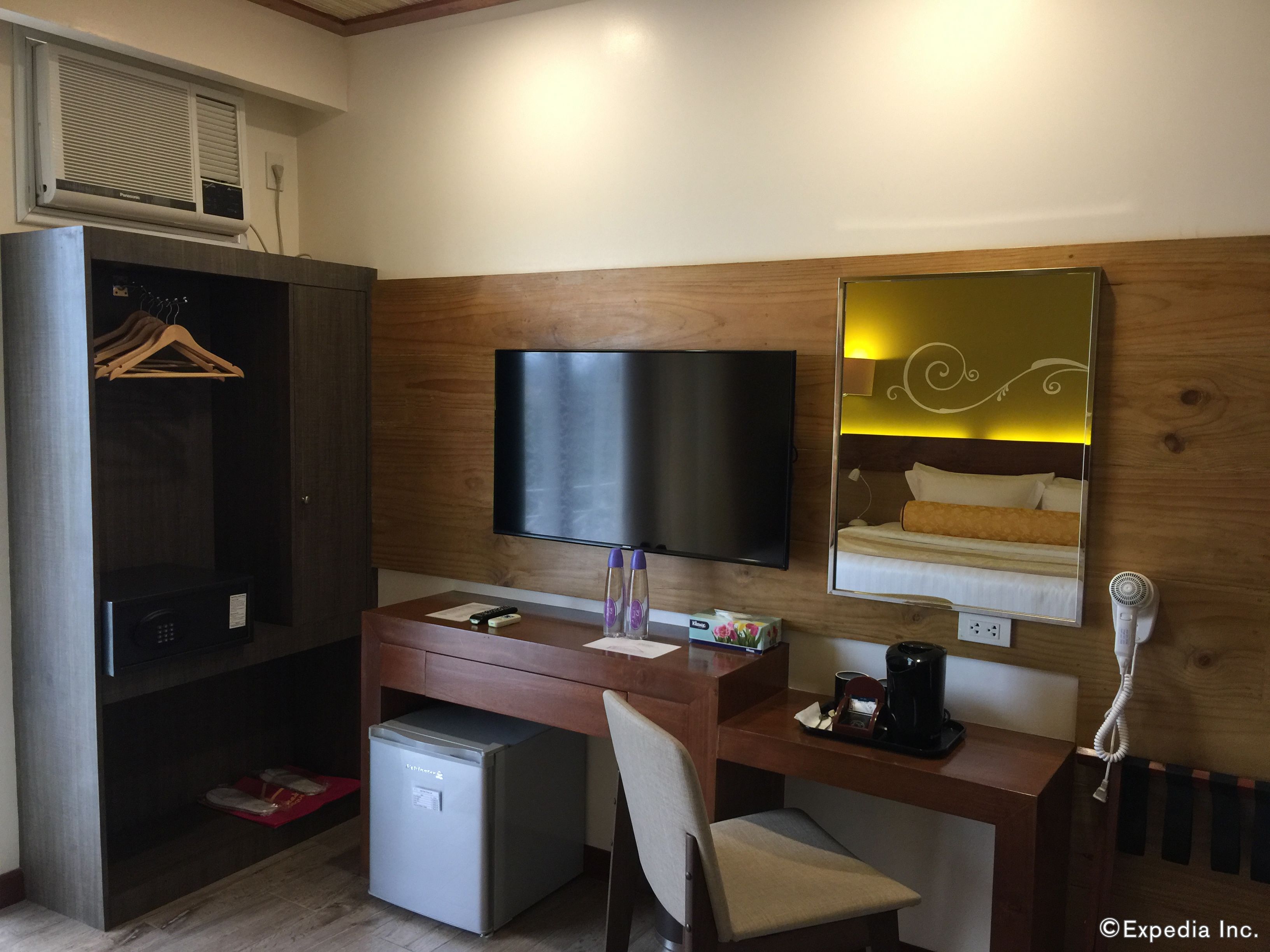 Une chambre d’hôtel équipée d’un bureau en bois, d’une télévision, d’un mini-réfrigérateur, d’une chaise, d’un lit avec une tête de lit et d’un miroir.