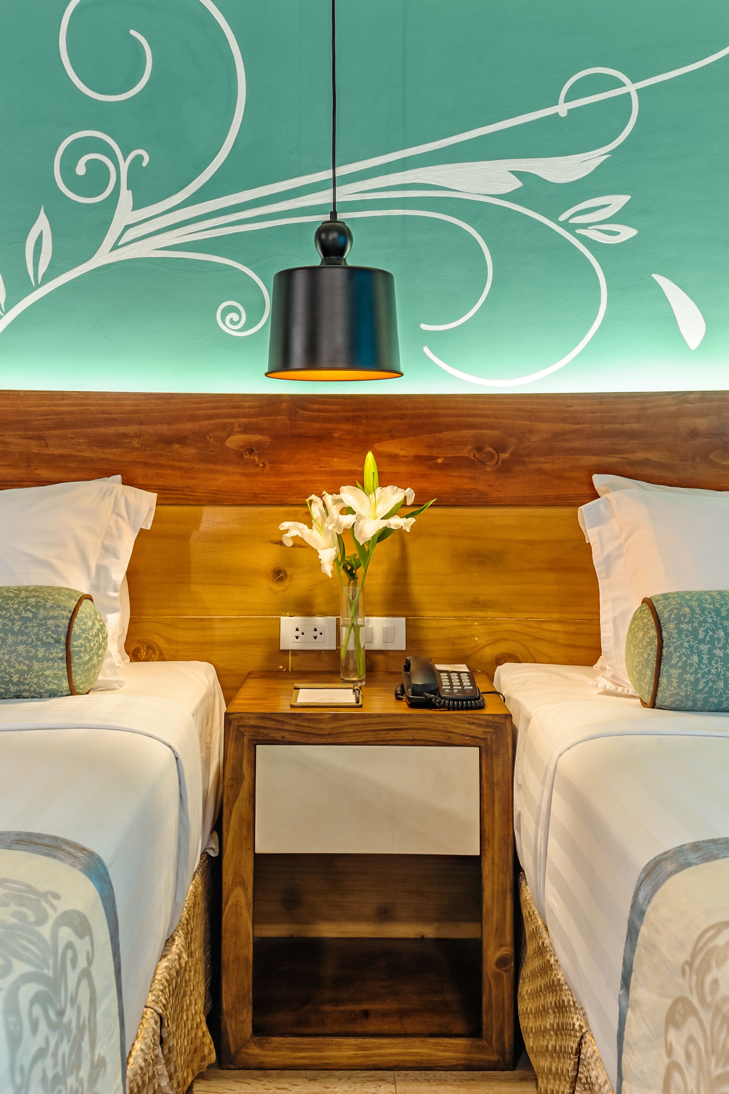 Une chambre d’hôtel avec deux lits, une tête de lit en bois, une table de chevet avec un téléphone, un vase avec des fleurs et un mur orné d’un motif décoratif.