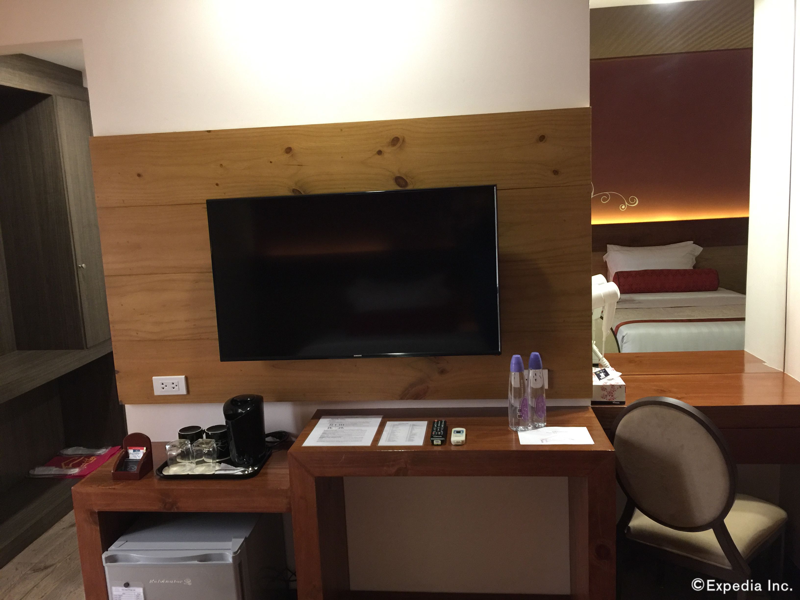 A hotel room with a wooden desk, a flat-screen TV, a bed with a red pillow, and a bedside table with a phone and a lamp.