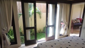 A bedroom with a bed, a sliding glass door leading to a balcony with plants, and a view of a dining area. - Praschita Bali (Denpasar)