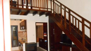 A spacious living area with a wooden staircase, a kitchen with a tiled backsplash, and a black entertainment unit. - Praschita Bali (Denpasar)