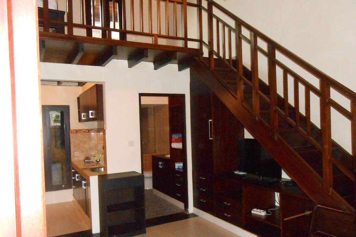 Amplia sala de estar con escalera de madera, cocina con salpicadero de azulejos y mueble de entretenimiento negro.