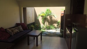 A living room with a sofa, a small table, and a kitchen area with cabinets and a refrigerator. - Praschita Bali (Denpasar)