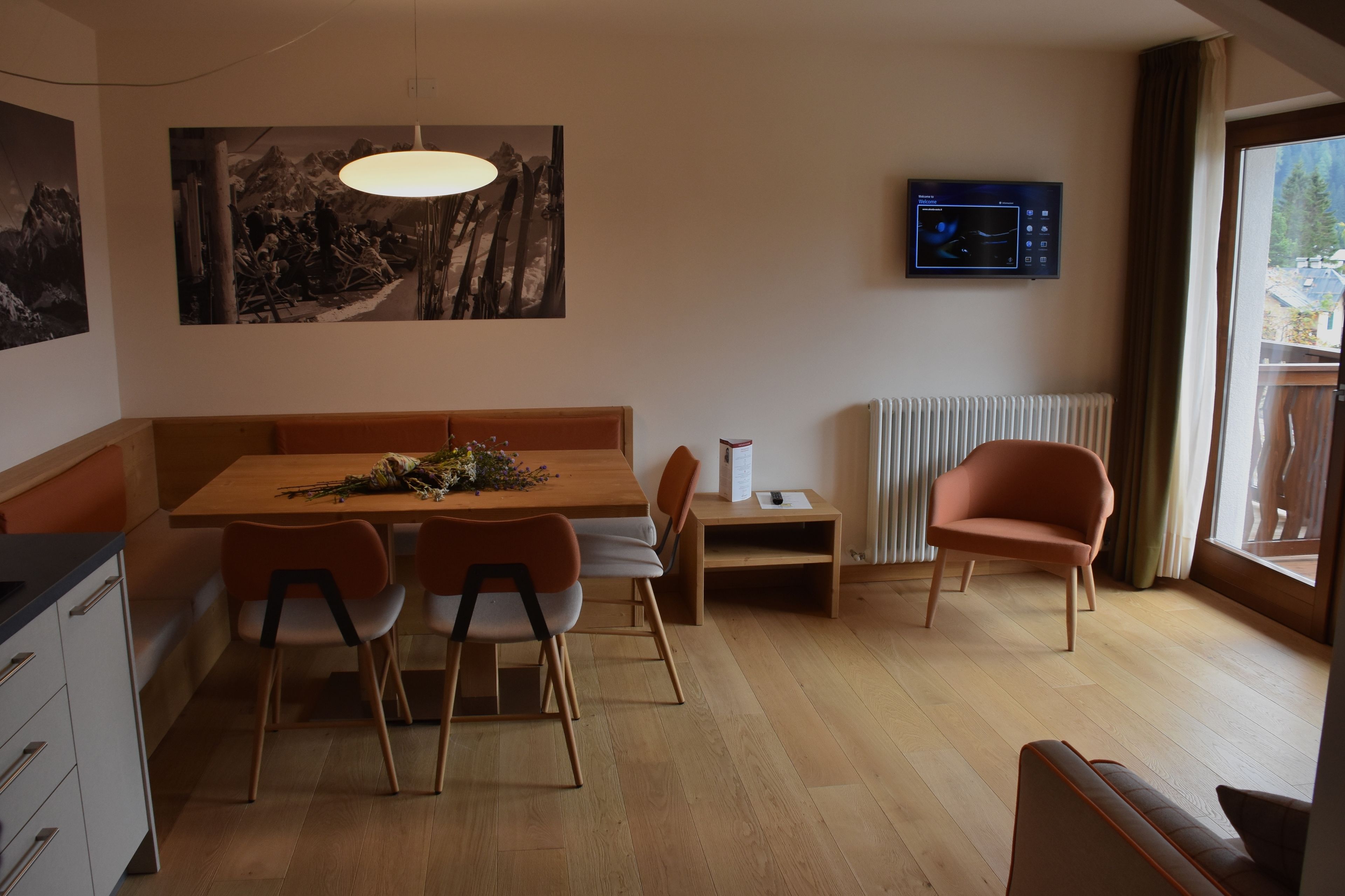 Un espace repas moderne avec une table en bois, quatre chaises, un banc, une suspension, un téléviseur à écran plat et une fenêtre donnant sur des arbres.
