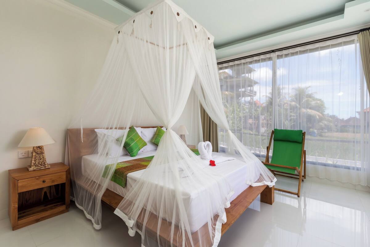 A neatly made bed with a canopy, a bedside table with a lamp, and a green chair by a large window.
