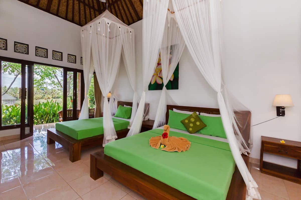 A room with two beds, each with a green bedspread and white canopy. There is a wooden bench, a nightstand with a lamp, and a wall-mounted light.