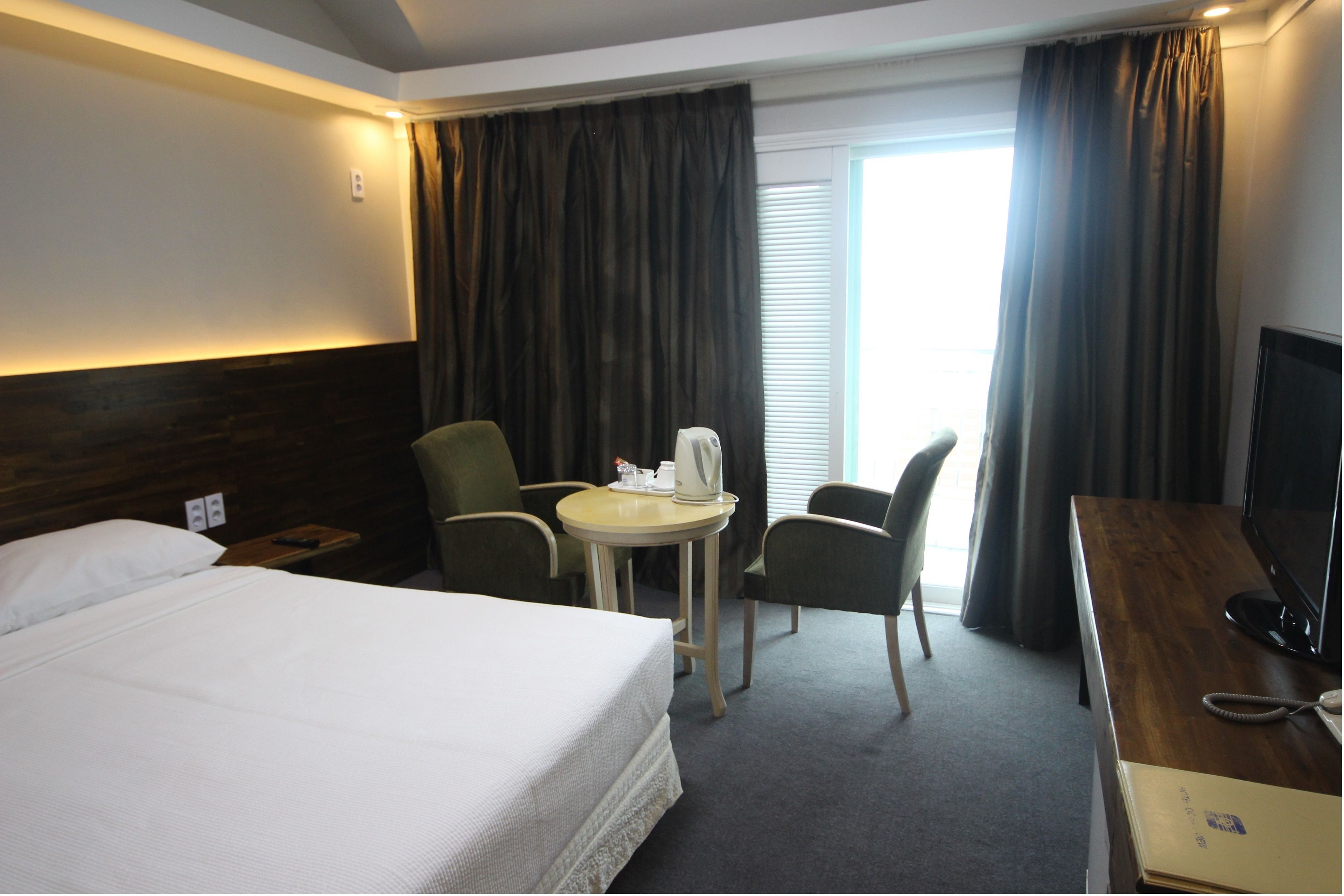 A hotel room with a large bed, a desk with a TV, a small table with a kettle, two chairs, and a window with curtains.