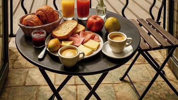 A balcony with breakfast items including croissants, bread, cheese, ham, apples, juice, and coffee, overlooking historic stone buildings and a cobblestone street.