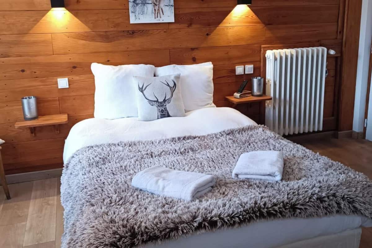 Makuuhuoneessa on puinen sängynpääty, valkoisilla vuodevaatteilla varustettu sänky, pehmeä matto ja kaksi seinälle asennettua lamppua.