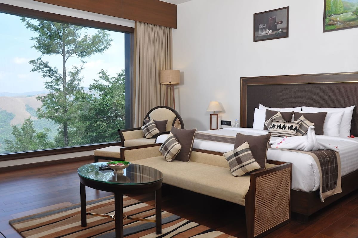 Ein Hotelzimmer mit Bett, Sofa und Tisch, große Fenster mit Blick auf Bäume und Hügel.