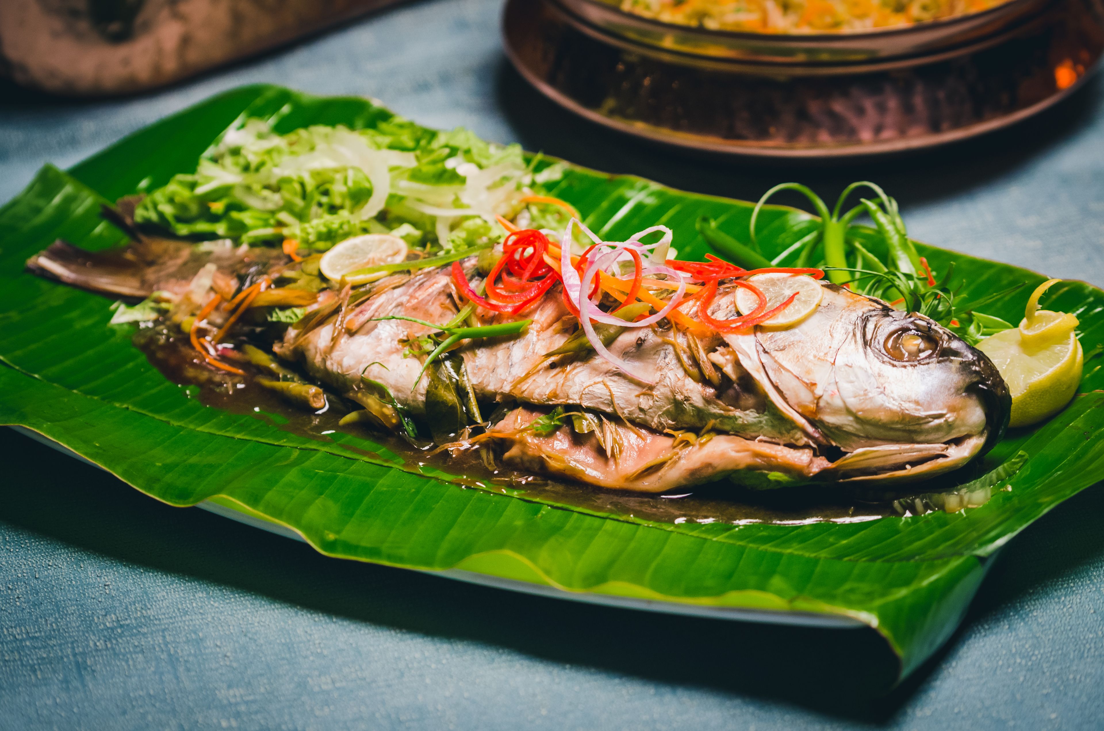 A whole fish on a banana leaf with garnishes and a bowl in the background.