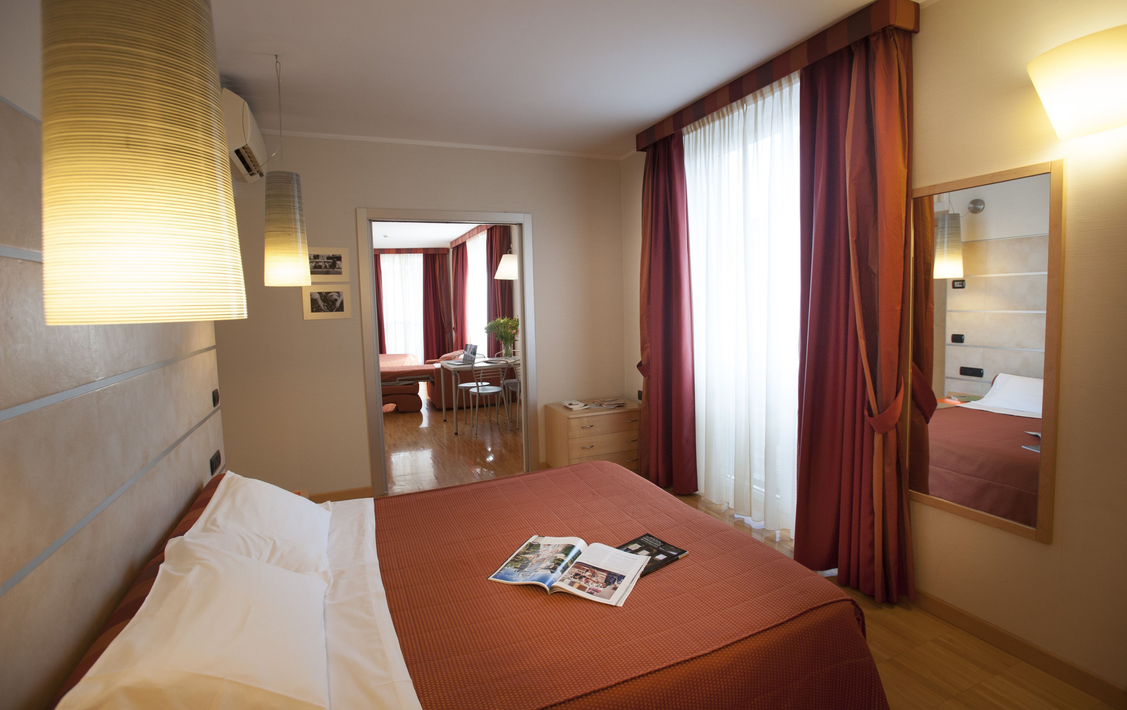 A hotel room with a bed, a desk, and a chair. There is a window with curtains, a lamp, and a mirror on the wall.