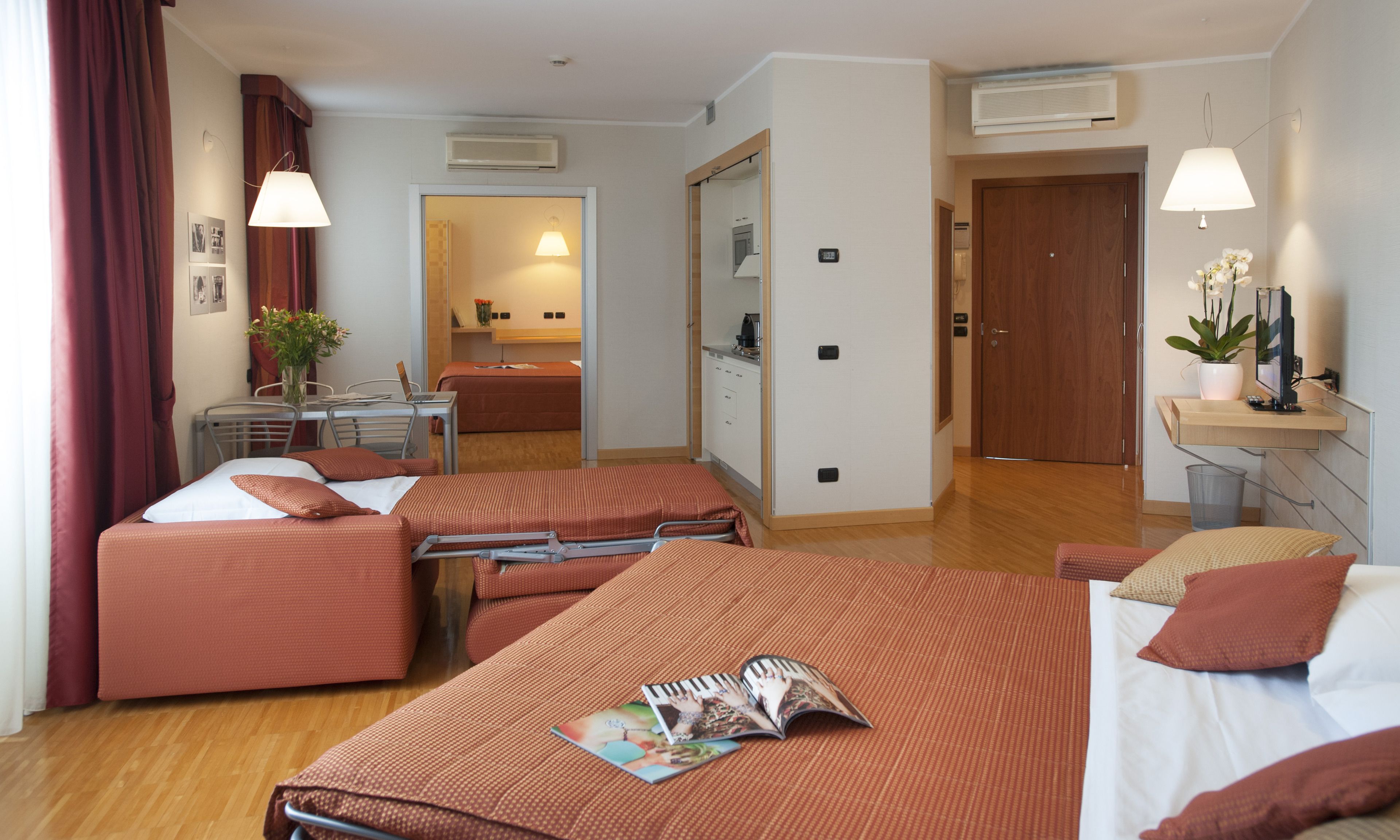 Una habitación de hotel con dos camas, un escritorio, una silla, una mesita, una planta en maceta y un espejo.