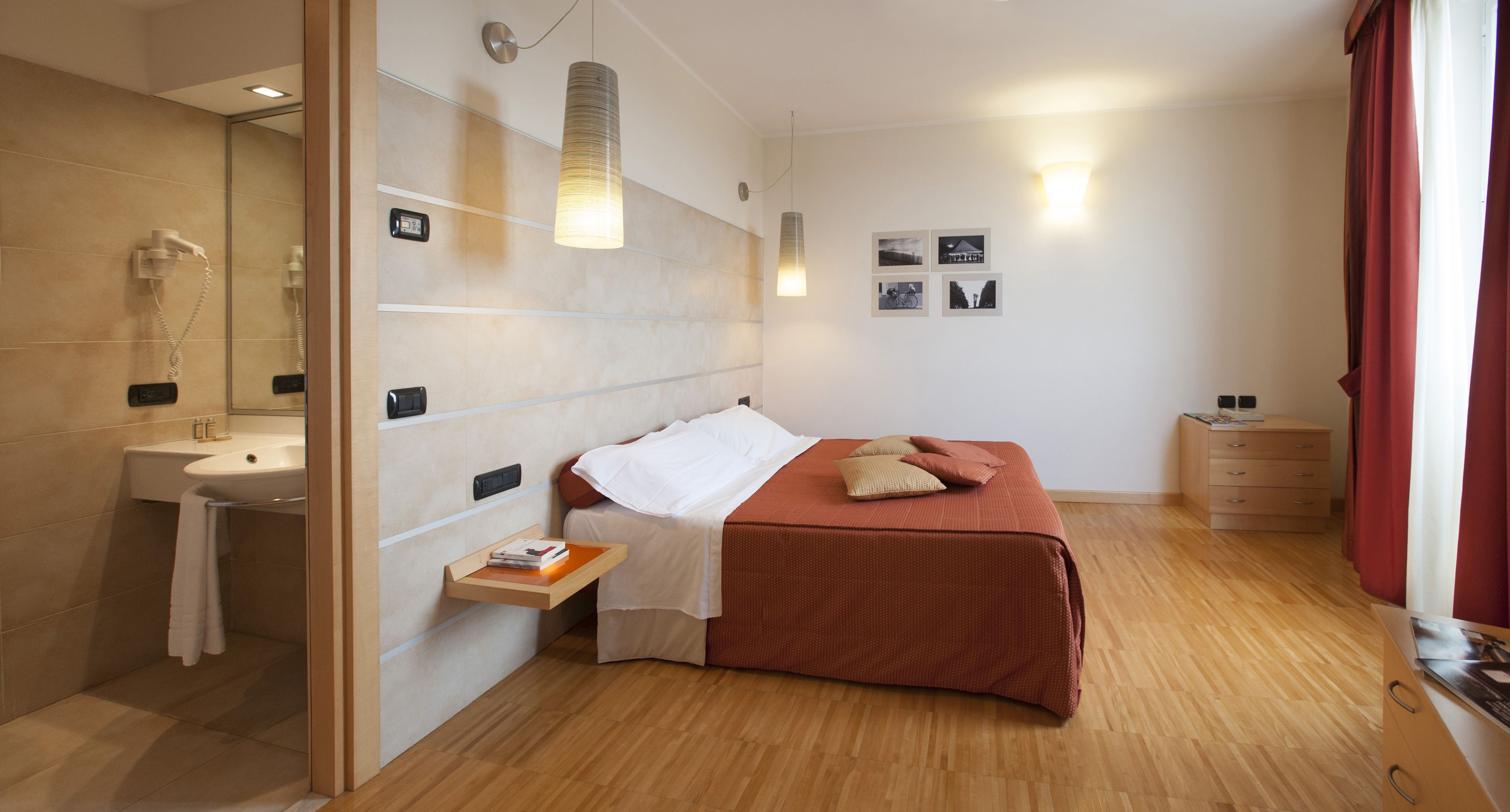 Un dormitorio moderno con piso de madera, una cama con cubierta roja, una mesita de noche con libros, una lámpara de pared y un estante pequeño con una toalla.