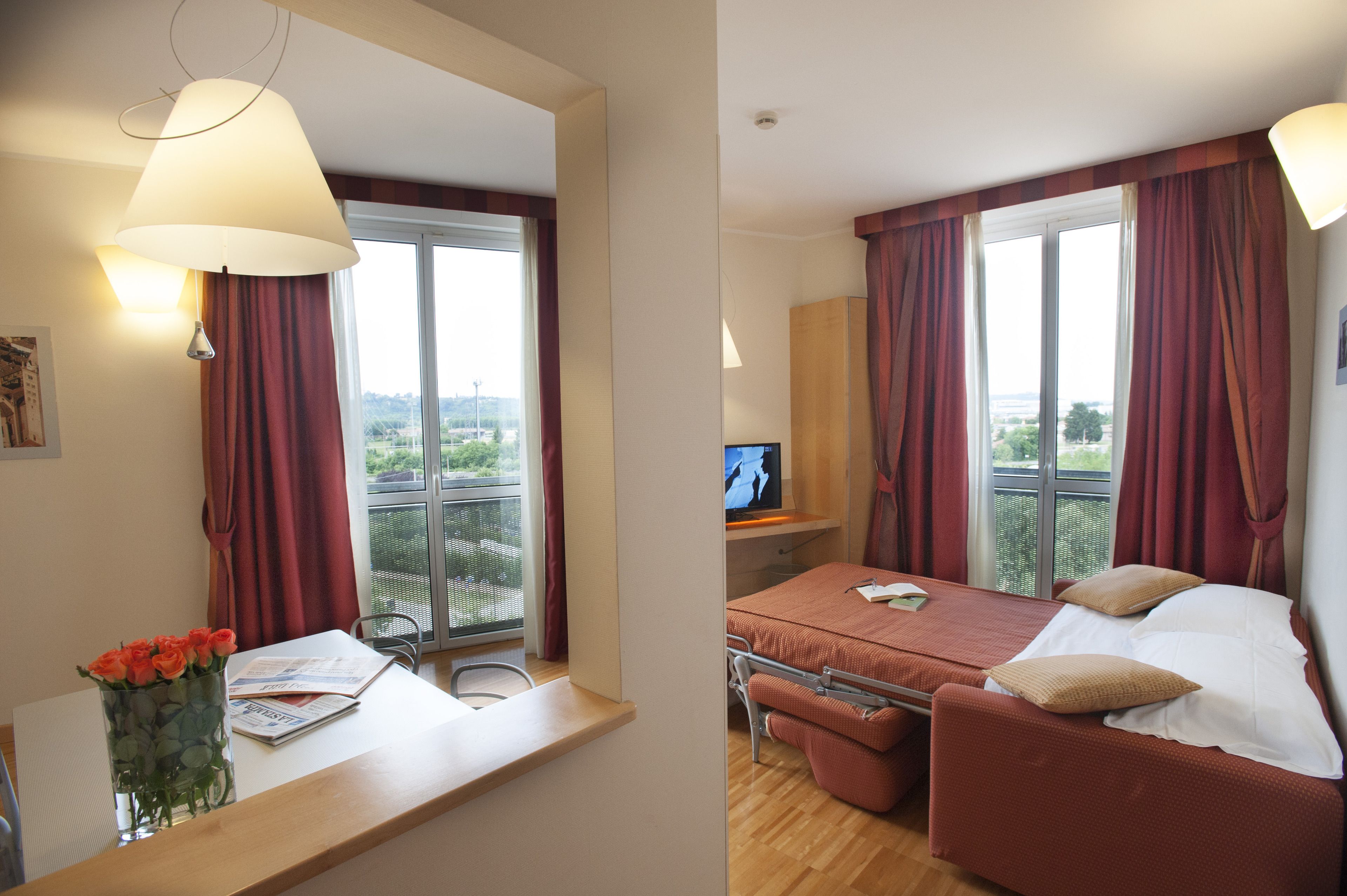 Une chambre d’hôtel avec un lit, un bureau avec un ordinateur, une fenêtre avec des rideaux et un vase avec des fleurs.