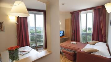 Une chambre d’hôtel avec un lit, un bureau avec un ordinateur, une fenêtre avec des rideaux et un vase avec des fleurs.