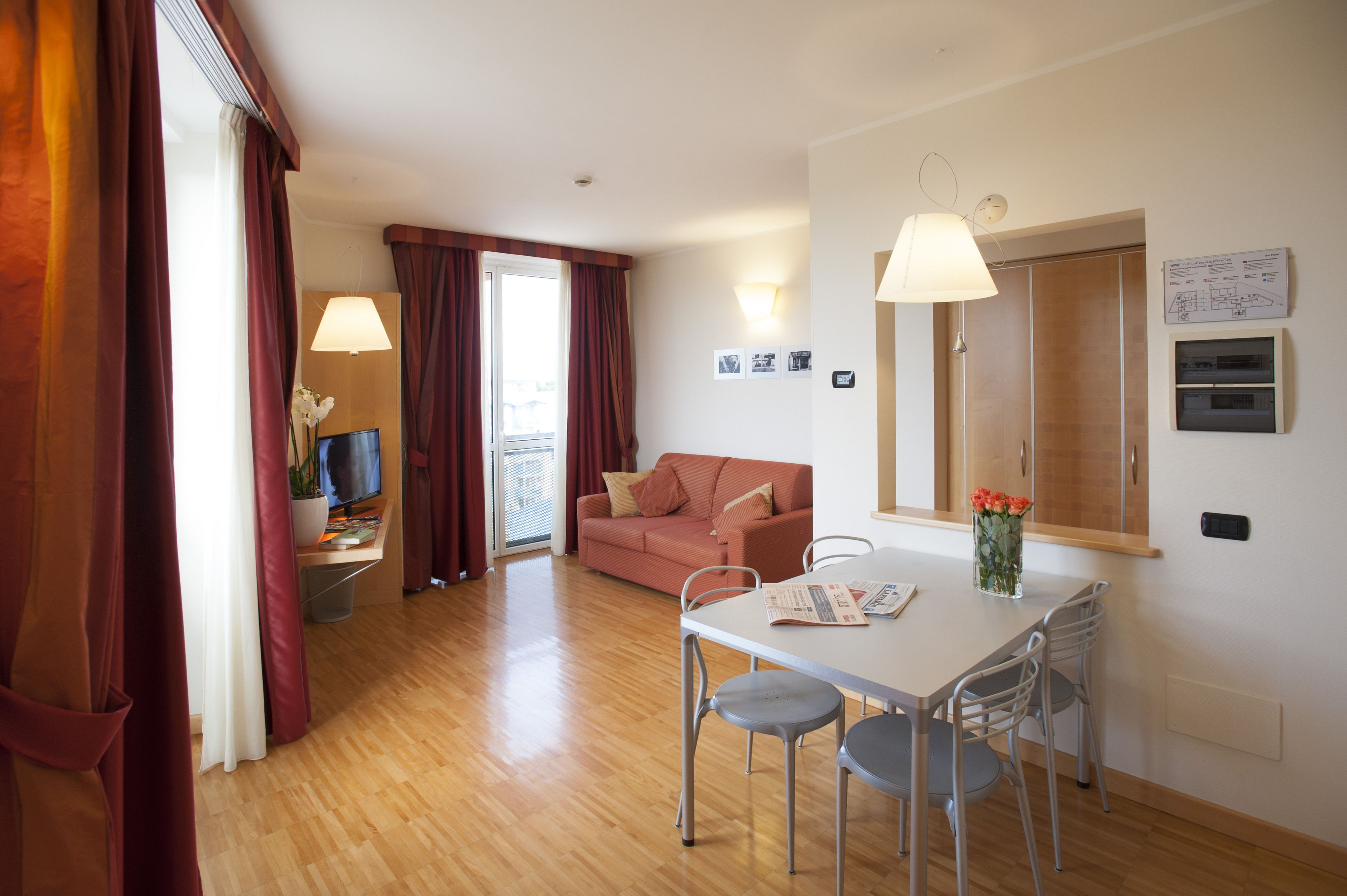 Ein modernes Hotelzimmer mit Holzboden, einem weißen Esstisch mit vier Stühlen, einem rosa Sofa, einem Fernseher und einem Fenster mit Vorhängen.