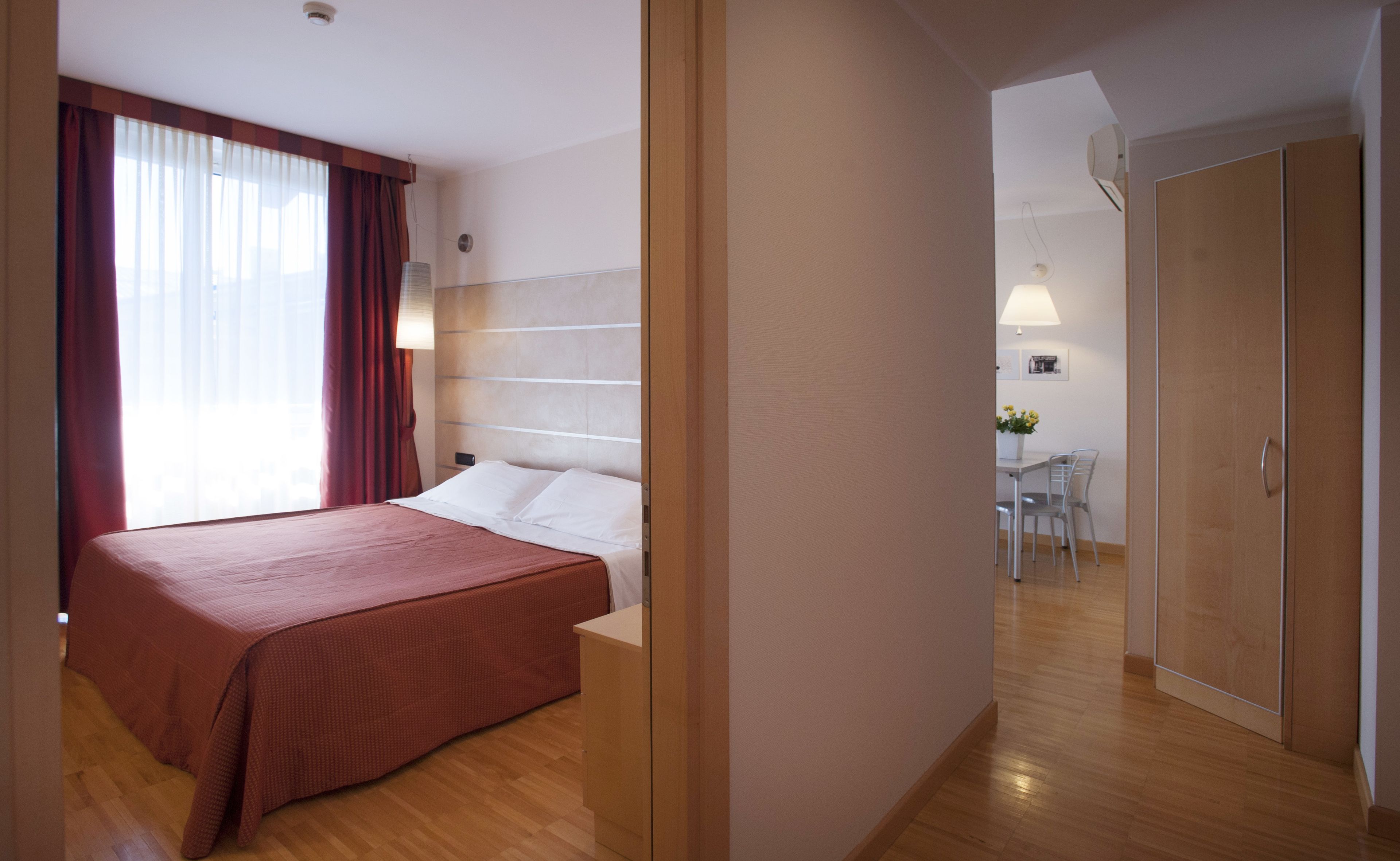 Een hotelkamer met een bed, een bureau en een stoel. Er is een deur die naar een andere kamer leidt.