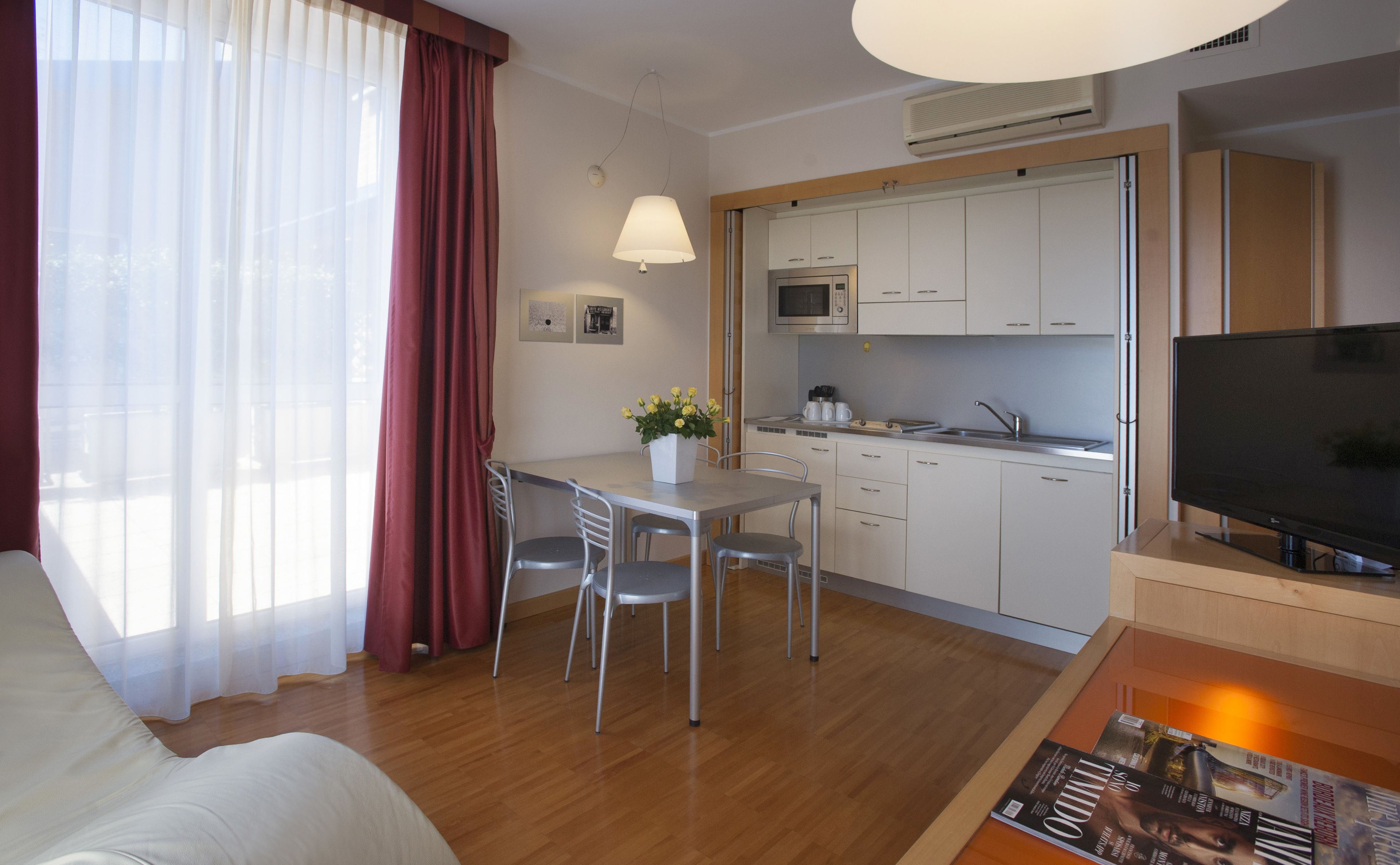 A modern hotel room with a dining area, kitchenette, and a flat-screen TV.