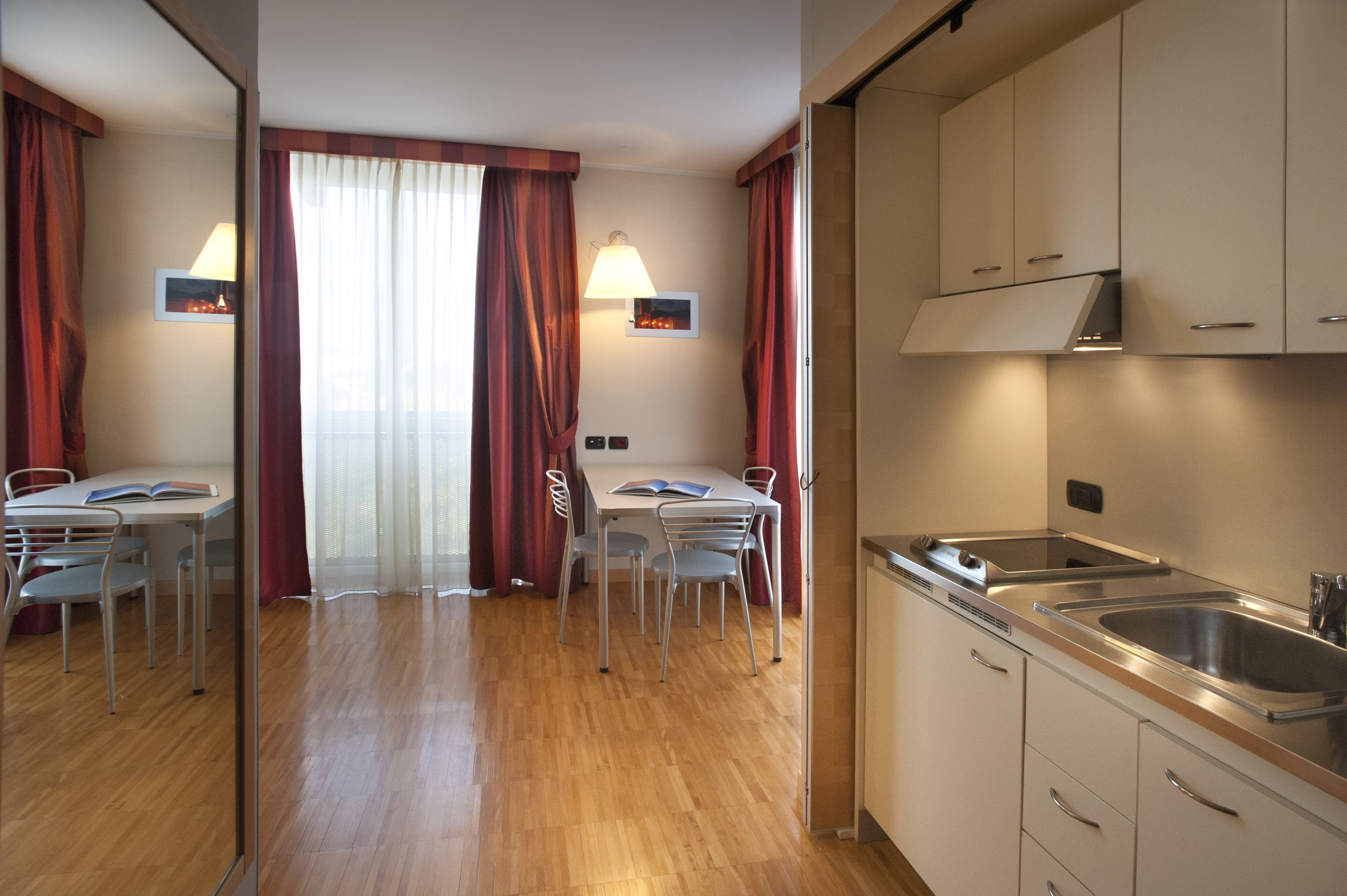 Habitación de hotel con suelo de madera, una mesa de comedor con sillas, un área de cocina con un lavabo y armarios, y una ventana con cortinas.