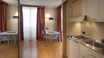 Habitación de hotel con suelo de madera, una mesa de comedor con sillas, un área de cocina con un lavabo y armarios, y una ventana con cortinas.
