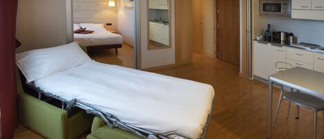 Quarto de hotel compacto com uma cama de solteiro, uma kitchenette e um banheiro visível através de uma porta aberta.