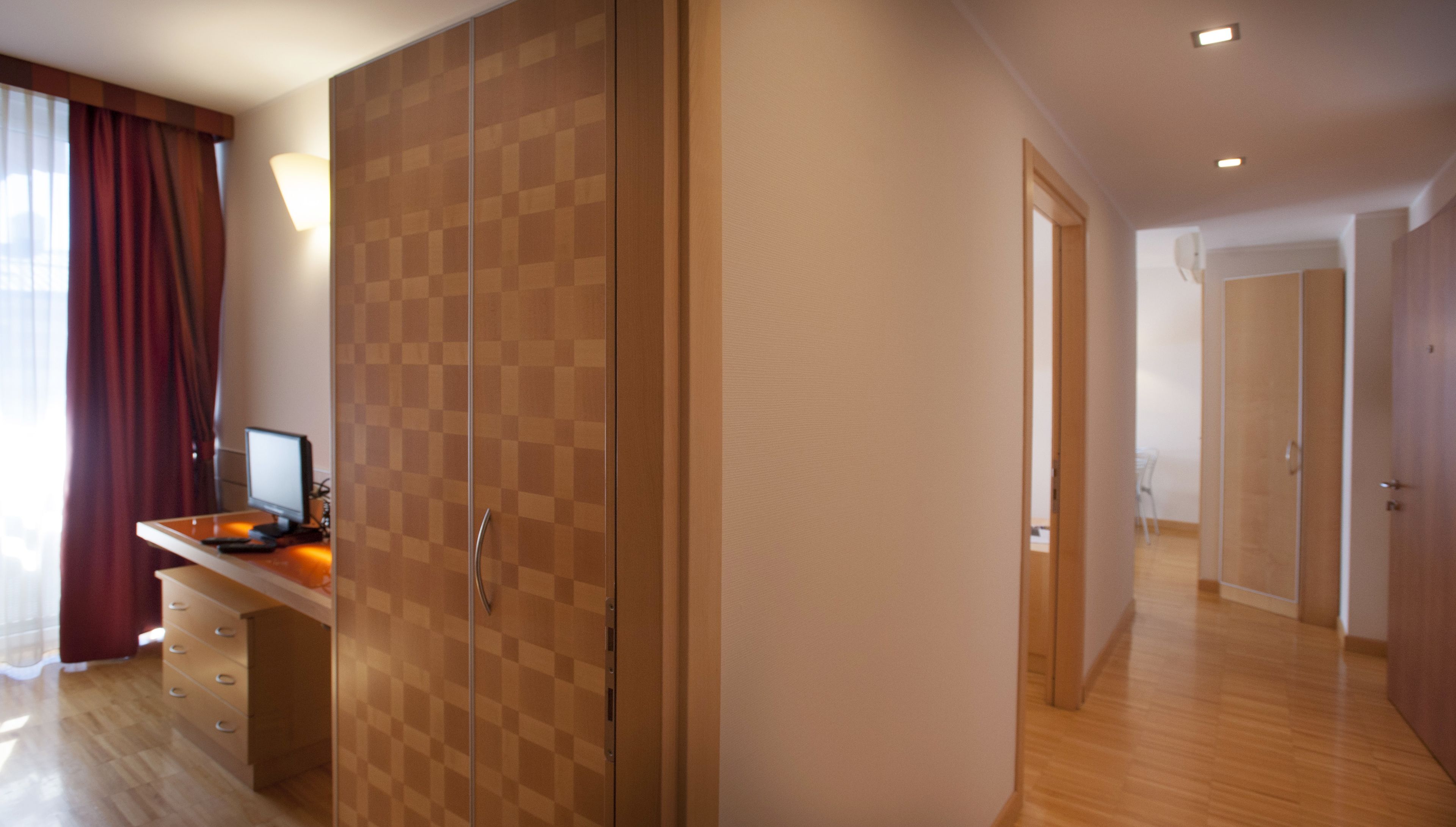 A modern hotel room with a large wardrobe, a desk with a computer, and a view of the hallway.