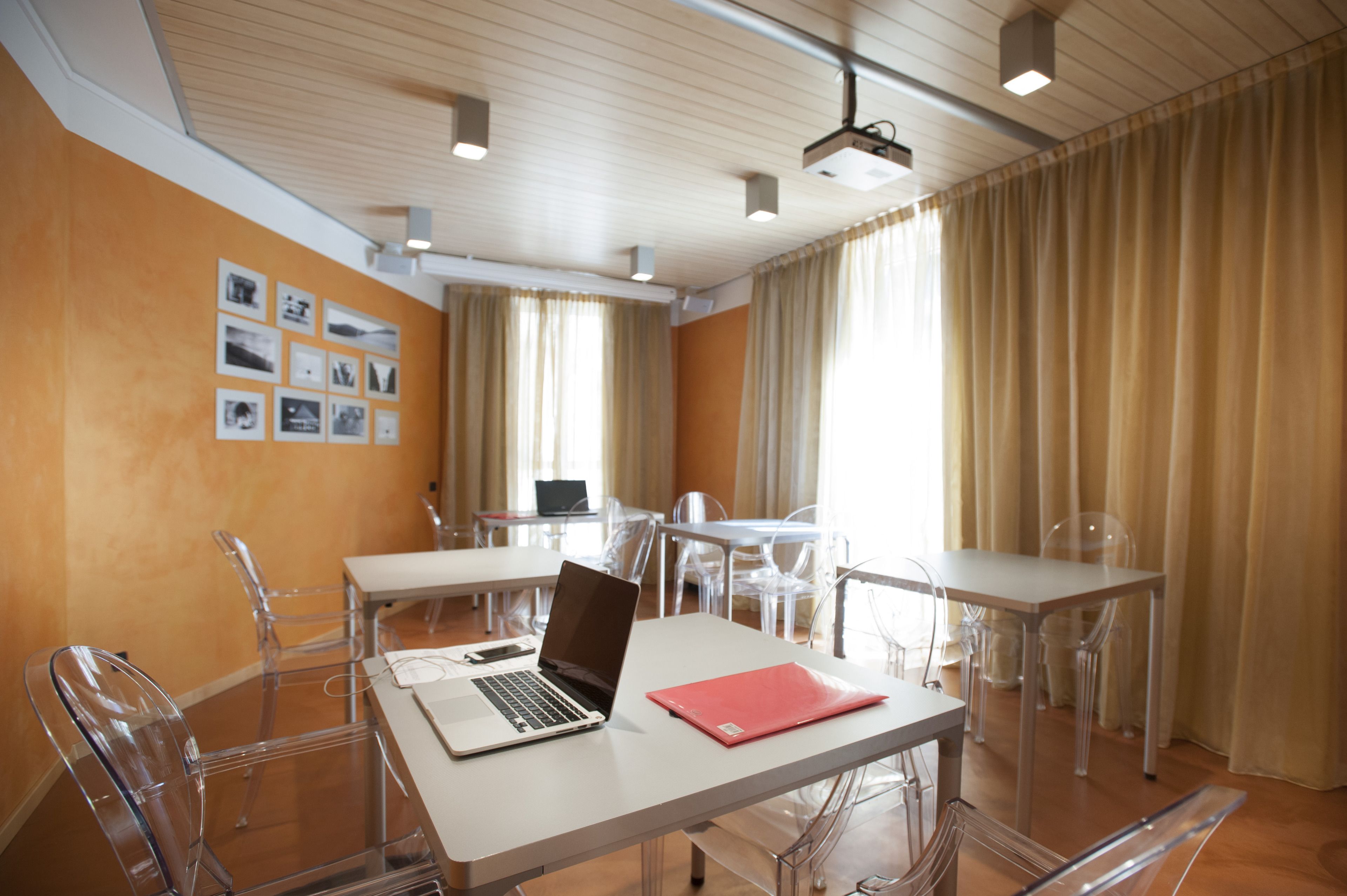 A conference room with a white table, a laptop, and a red folder. There are clear plastic chairs around the table. The room has beige curtains and a projector on the ceiling.